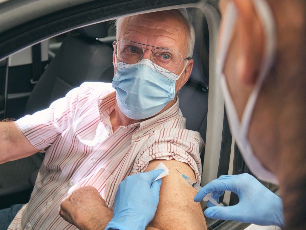 γυναίκα γιατρός με χειρουργική ενδυμασία κρατώντας σύριγγα εμβολιάζει ηλικιωμένο άντρα εντός του αυτοκινήτου του εν μέσω της πανδημίας του Coronavirus